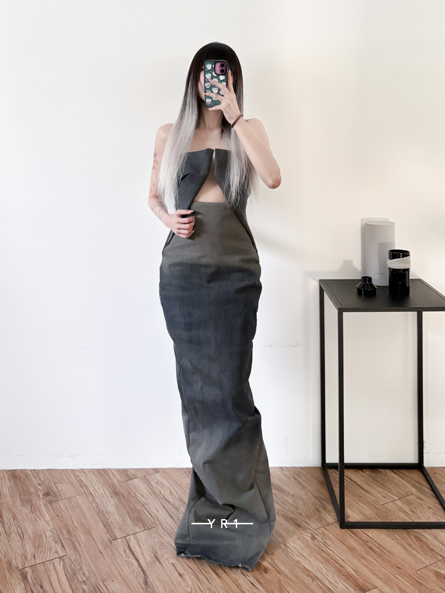 Model taking mirror selfie wearing a long, dark gray, avant-garde designer gown with cut-out details in a minimal room with wooden floor and black side table